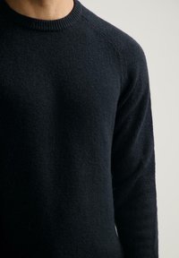 Nahaufnahme einer Person, die einen dunkelblauen Strickpullover mit Rundhalsausschnitt vor einem einfarbigen hellen Hintergrund trägt.