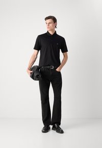 Black polo shirt with a collar and buttons, paired with black pants and shoes. Model holds a black bag, against a neutral background.
