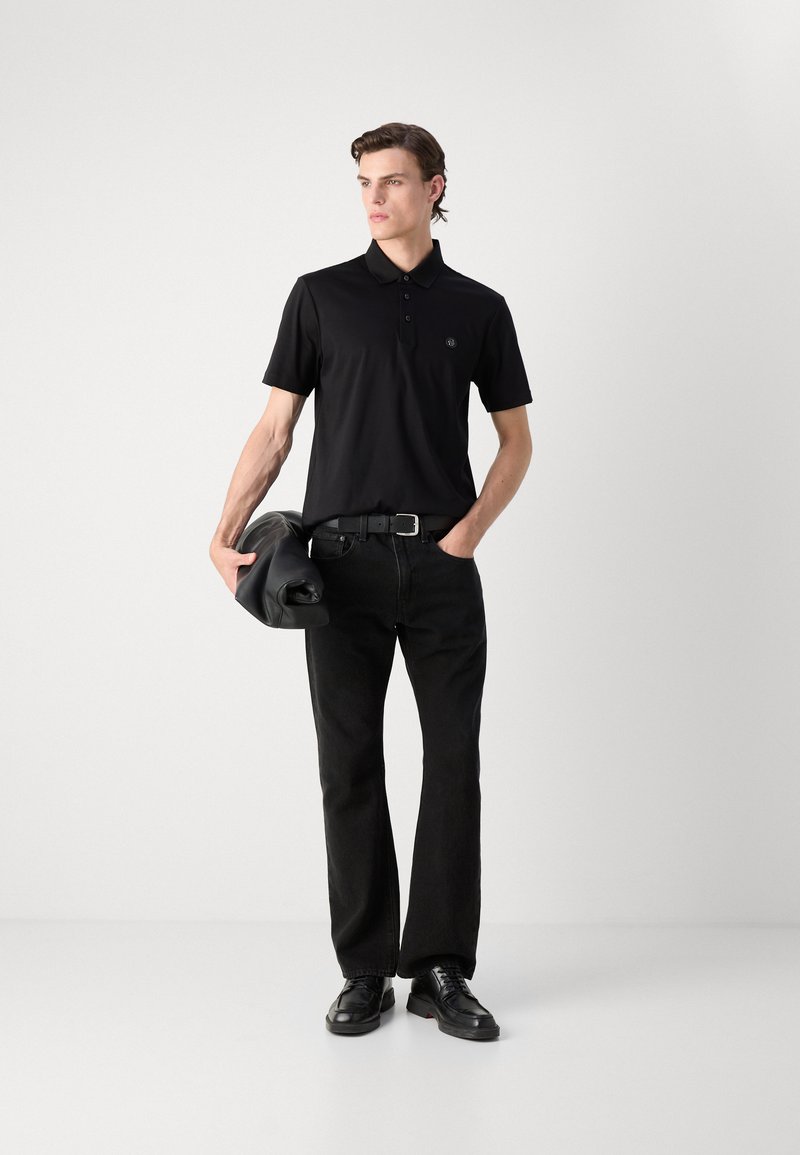 Schwarzes Poloshirt mit Kragen und Knöpfen, kombiniert mit schwarzen Hosen und Schuhen. Das Modell hält eine schwarze Tasche und steht vor einem neutralen Hintergrund.