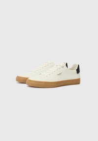 Zapatillas de lona en color crema con suela de goma, que presentan un acento negro en el talón y seis ojales para los cordones. Logo mostrado en el lateral.