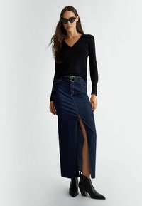 Zwarte lange mouwen top met een V-hals, gecombineerd met een donkere denim maxi rok met een split aan de voorkant en een zilveren gesp riem. Zwarte enkelhoge laarzen met hak.