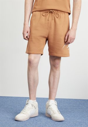 Personne portant un short beige à cordon avec un motif blanc sur la jambe gauche et des baskets blanches montantes, debout sur un tapis bleu.