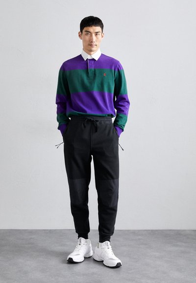 Camisa polo riscada em verde e roxo com colarinho branco, combinada com calças de jogging pretas com painéis e ténis brancos.