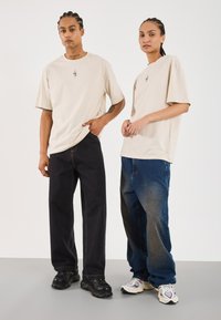 Beige übergroße T-Shirts mit einem kleinen Grafikdruck, kombiniert mit dunkelblauen und hellblauen baggy Jeans und klobigen Sneakers in Schwarz und Weiß.