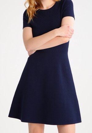 Žena v texturovaných tmavě modrých krátkých šatech s přiléhavým živůtkem a rozšířenou sukní, s rukama založenýma v pase.