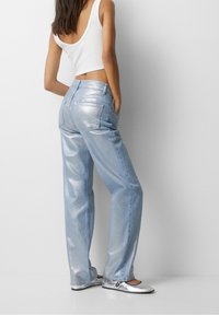 Vaqueros metálicos de color azul claro con un corte relajado, que cuentan con bolsillos traseros y una cintura de altura media, combinados con zapatos planos plateados y una camiseta sin mangas blanca.