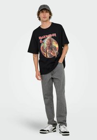 Camiseta de algodón negra con un gráfico colorido de Iron Maiden, combinada con jeans grises y zapatillas en blanco y negro. Gorro ajustable en la cabeza.