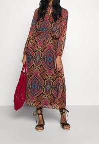 Robe longue à motifs avec un fond sombre et des tourbillons colorés ainsi que des motifs paisley. Accessoirisée avec un sac en crochet rouge et des sandales noires.