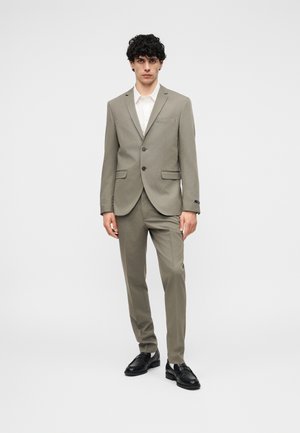 Hombre de pie vistiendo un traje gris claro con blazer de dos botones, camisa blanca, pantalones a juego y zapatos de vestir negros contra un fondo liso.