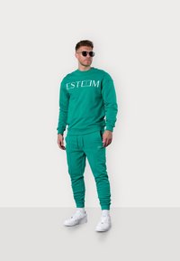 Türkiser Sweatshirt mit dem weißen "ESTEEM"-Logo, kombiniert mit passenden Jogginghosen. Beide aus einem weichen Stoff gefertigt. Weiße Sneakers runden den Look ab.