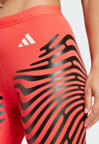 Red athletic leggings with a smooth texture, featuring a black wavy pattern and white "adizero" logo at the waistband.