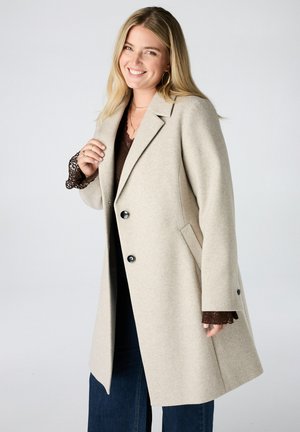 Femme souriante aux longs cheveux blonds portant un manteau en laine beige par-dessus un haut en dentelle marron et un jean bleu foncé sur un fond clair uni.