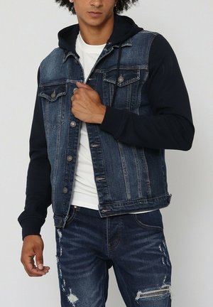 Veste en denim avec des manches bleu foncé, des poches sur le devant et des boutons en métal. Portée sur une chemise blanche, associée à un jean bleu déchiré.