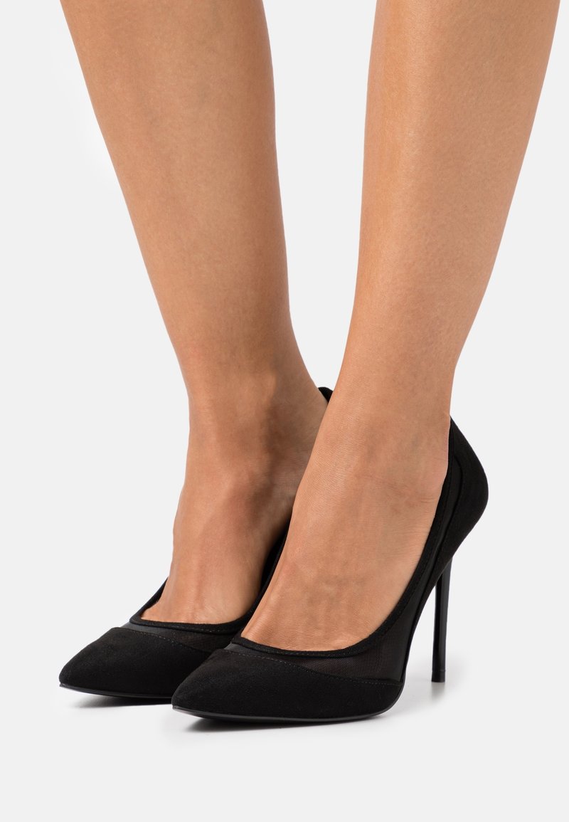 Pieds d'une femme portant des escarpins noirs à bout pointu sur fond blanc uni.
