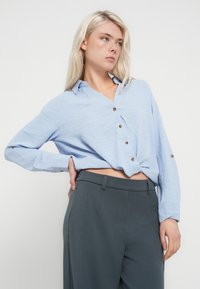Jeune femme aux longs cheveux blonds portant une chemise bleu clair boutonnée et un pantalon taille haute gris foncé, posant avec une main sur la hanche.