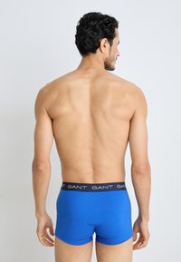 Modré bavlnené boxerky s čiernym pásom s opakujúcim sa logom "GANT", zobrazené zozadu na holom trupe.