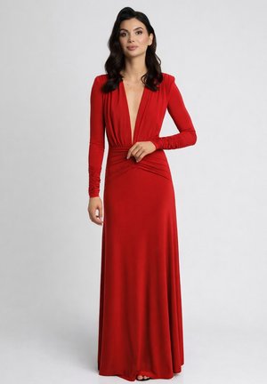 Kobieta o ciemnych włosach ubranа w długą, czerwoną suknię z głębokim dekoltem w kształcie litery V i długimi marszczonymi rękawami, stojąca na jednolitym jasnym tle.
