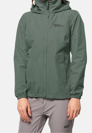 Nainen, joka pukeutunut vihreään vedenpitävään takkiin, jossa on huppu ja joustavat hihansuut, yhdistettyinä vaaleanharmaisiin housuihin, molemmat merkkiltään Jack Wolfskin.