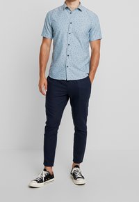 Lichtblauwe overhemd met korte mouwen en kleine donkere geometrische patronen, gedragen met marineblauwe broek en zwarte sneakers.