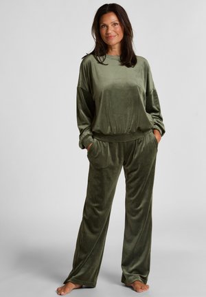 Frau steht barfuß und trägt ein olivegrünes Velour-Langarmshirt und passende weite Hosen mit den Händen in den Taschen.