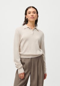 Suéter de polo tricotado em bege com colarinho, mangas longas e bainha canelada. Usado com calças pregueadas em castanho claro, apresentando uma textura suave.