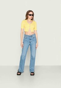 Top amarelo cropped com mangas curtas e fecho de botão, combinado com jeans claros de cintura alta e sandálias pretas de plataforma.