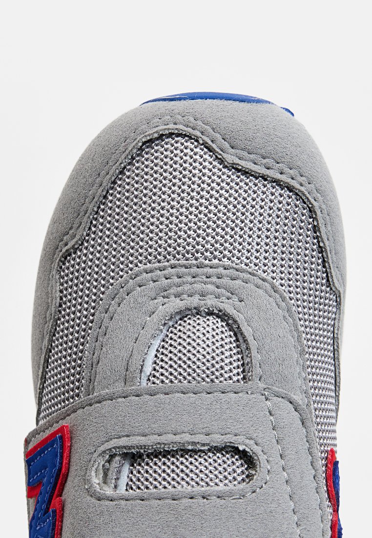 Chaussure de sport grise avec une tige en daim et en mesh, une sangle Velcro, et des accents rouges et bleus, avec un bout arrondi et une surface texturée.