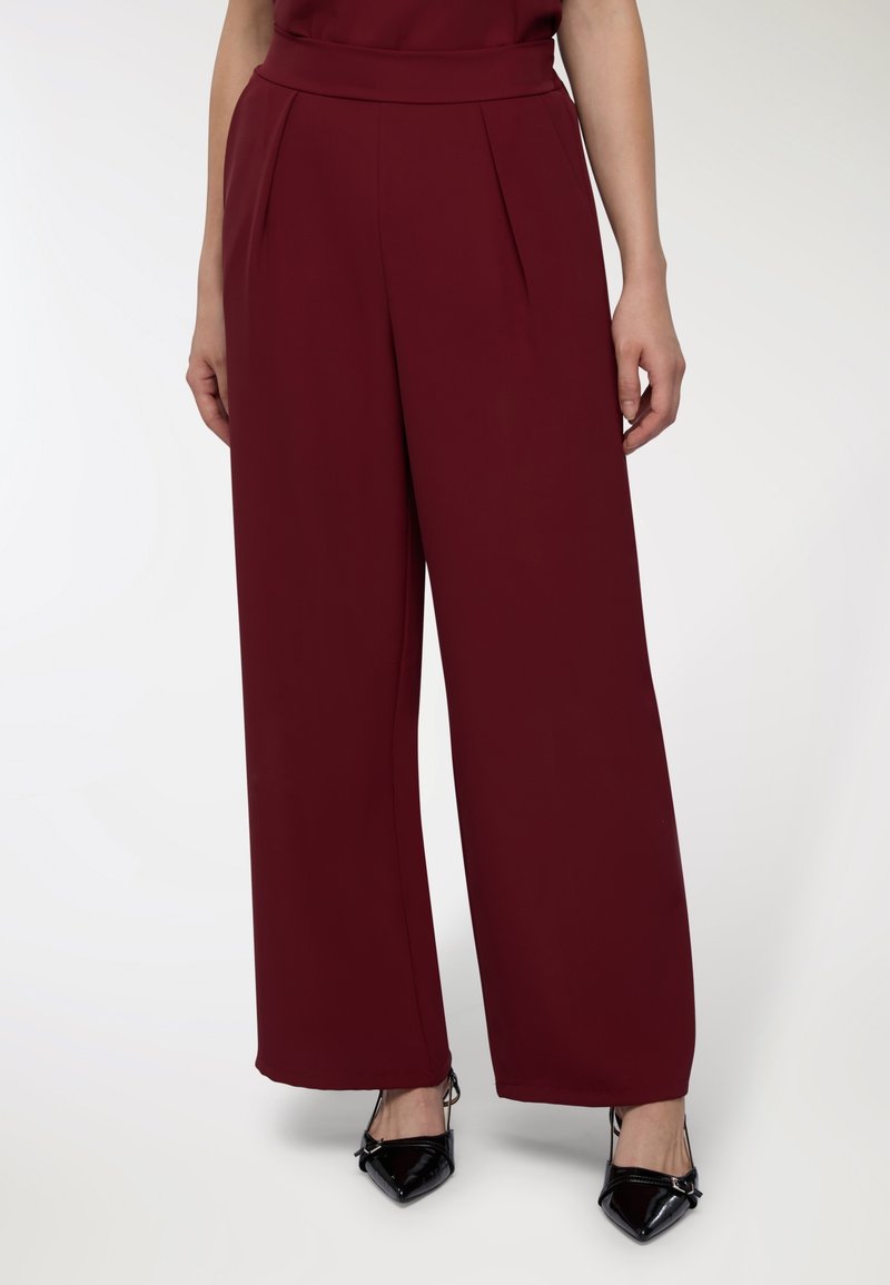 Pantaloni ampi bordeaux con vita alta, caratterizzati da pince e una texture liscia, abbinati a tacchi a punta neri.