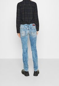 Sininen haalistu denim-housut, joissa on kapea leikkaus. Takana on taskut, joissa on ompeluyksityiskohtia, ja vyötäröllä on erottuva muotoilu.