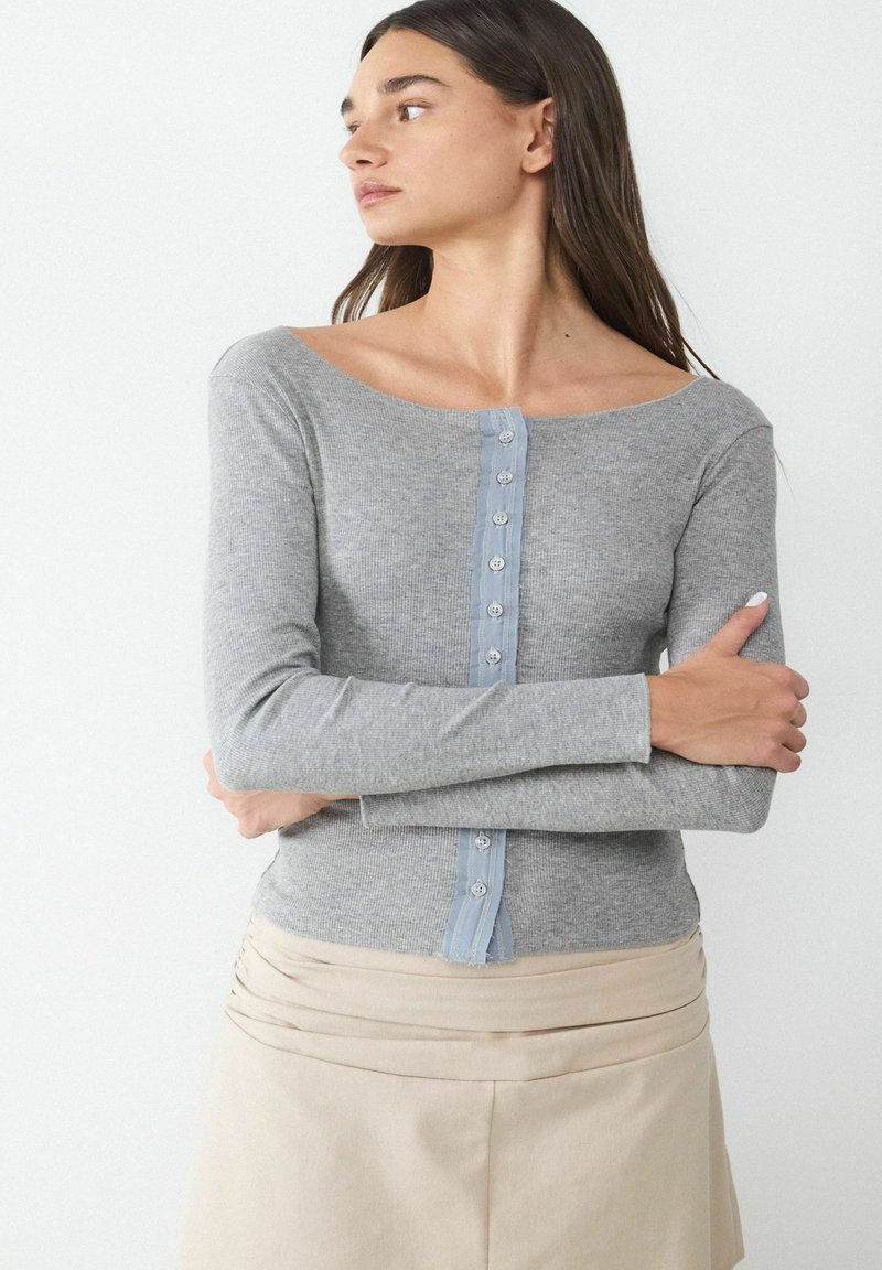 Gray long-sleeve shirt with a ribbed texture, boat neckline, and light blue button placket down the front. Worn with a beige skirt.