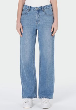 Vue de face d’une personne portant un jean bleu clair à jambes larges avec un bouton et une fermeture éclair, associé à des baskets blanches et un haut blanc.