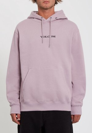Mand iført lys lilla Volcom hættetrøje med forlommes lomme og snøre i hætten, sorte bukser, mod ensfarvet baggrund.