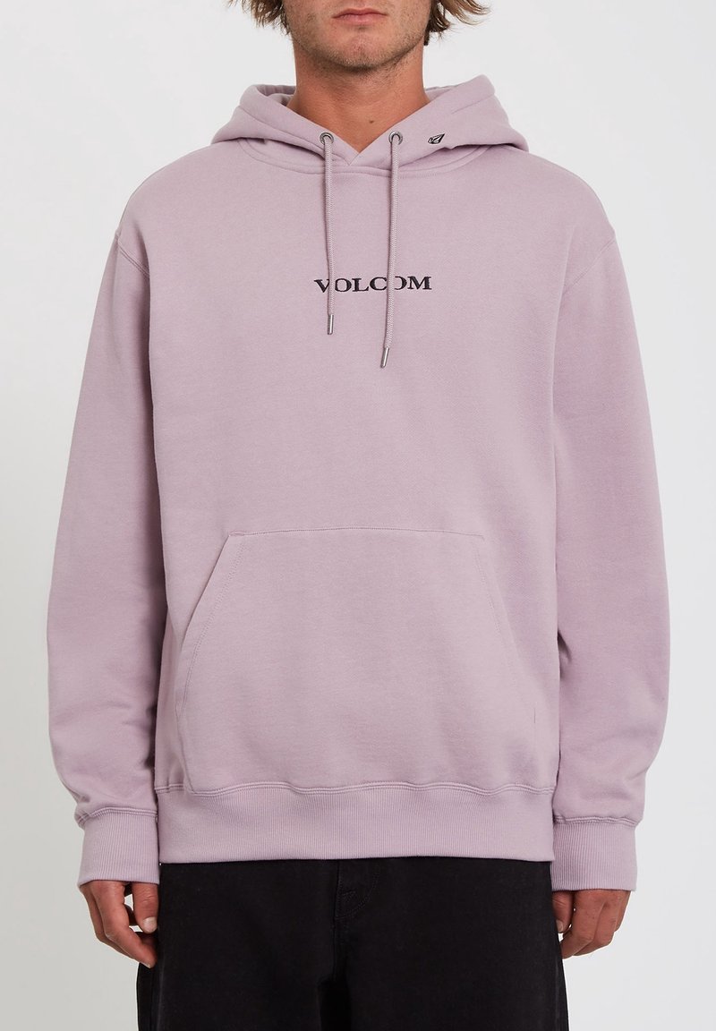 Homme portant un sweat à capuche Volcom violet clair avec poche kangourou devant et capuche à cordon, pantalon noir, sur fond uni.