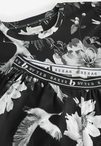 Čierne šaty s kvetinovým vzorom, obsahujúce žeriavy a kvety. Okrúhly výstrih a elastický pás s brandovaním "By Ted Baker".
