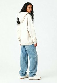 Felpa oversized beige chiaro con cappuccio regolabile, abbinata a jeans in denim azzurro larghi e sneakers bianche.