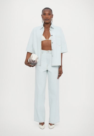 Camisa de denim azul-claro e jeans de perna larga, combinados com um top de bikini branco e saltos brancos modernos. Os acessórios incluem uma clutch estruturada.