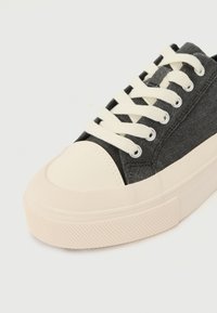 Schwarze Sneaker aus Canvas mit strukturiertem Design, Gummizehenkappe und cremefarbenen Schnürsenkeln. Verfügt über eine dicke, gemusterte Sohle für besseren Halt.