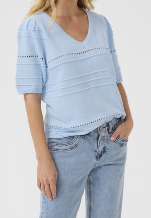 Femme portant un haut tricoté bleu clair à manches courtes et col en V avec des lignes horizontales texturées et un jean en denim bleu clair, main dans la poche.