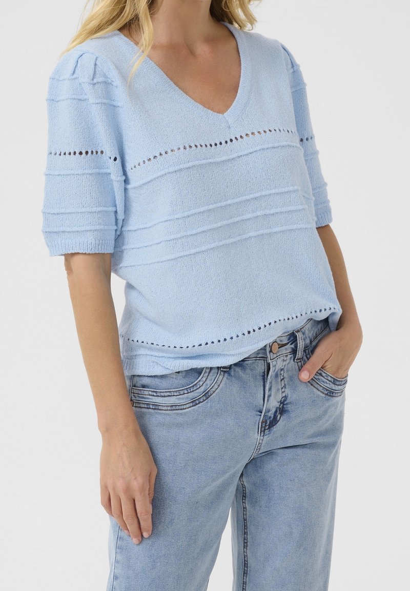 Femme portant un haut tricoté bleu clair à manches courtes et col en V avec des lignes horizontales texturées et un jean en denim bleu clair, main dans la poche.