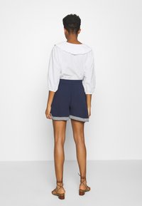 Blouse blanche à manches longues avec col à volants associée à un short bleu marine avec un ourlet décoratif. Mannequin portant des sandales à talons marron.