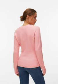 Femme aux cheveux bruns attachés en chignon, portant des boucles d'oreilles argentées en forme d'anneau, un pull rose à manches longues et un jean bleu, vue de profil et regardant par-dessus son épaule.