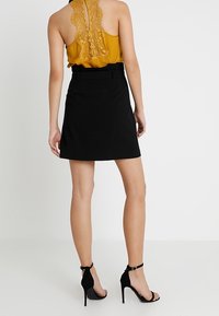 Gonna nera con vita aderente e passanti per cintura, abbinata a una maglia gialla senape con texture e dettagli in pizzo sulla schiena. Sandali neri.