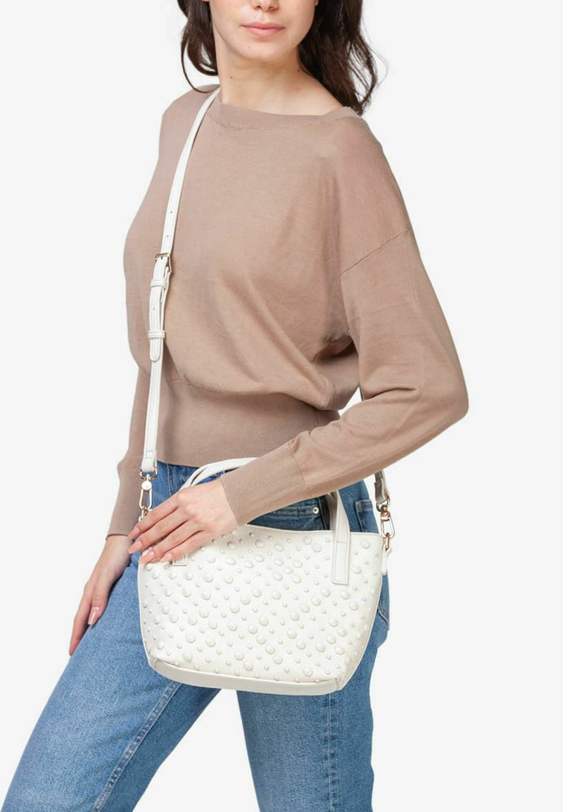 Hvid håndtaske med en struktureret, naglet overflade, som har en lang, justerbar strop. Sammen med en lysebrun sweater og blå jeans.