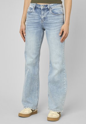 Personne portant un jean droit taille haute bleu clair avec poches avant et des baskets beiges avec semelles en caoutchouc, debout devant un fond gris.
