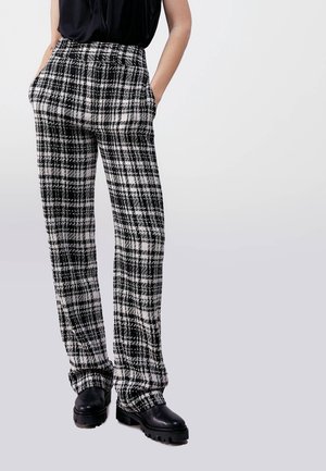 Person iført sorte og hvide ternede bukser med lige ben, med hænderne i lommerne og sorte læderstøvler med kraftig sål.