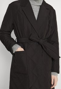 Černý prošívaný kabát s páskem v pase, má klopový límec a dvě boční kapsy. Pod ním je vidět šedý žebrovaný svetr.