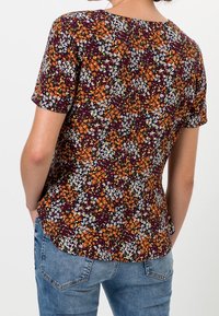 Blusa floral con fondo marrón, que presenta pequeñas flores naranjas, blancas y verdes. Mangas cortas, escote redondeado, silueta ajustada.