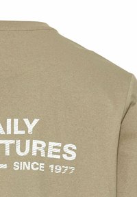 Beige bomulds t-shirt med korte ærmer, med hvid tekst: "DAILY ADVENTURES SINCE 1977" trykt i en fed, slidt skrift.