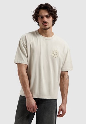 Man met krullend haar die een beige oversized T-shirt draagt met een gestructureerd embleem op de borst en donkergrijze spijkerbroek, staand tegen een effen achtergrond.