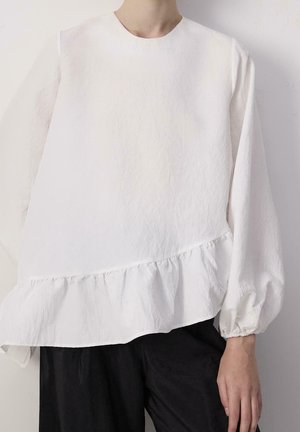 Personne portant un chemisier blanc à manches longues avec un ourlet volanté et des poignets élastiques, associé à un pantalon noir ample.